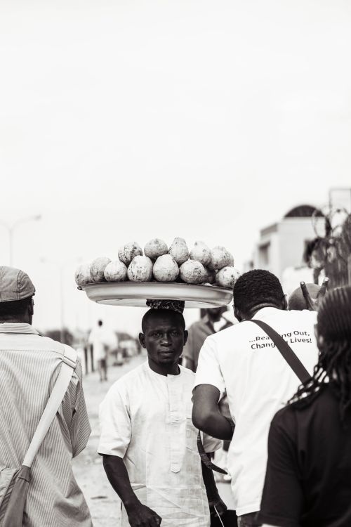 Free Black and white photo of a street vendor carrying fruits on his head in a busy market scene. Stock Photo