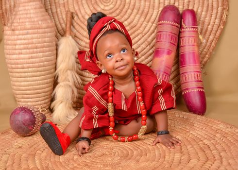Charming baby in vibrant red traditional African dress with jewelry on woven mat background.