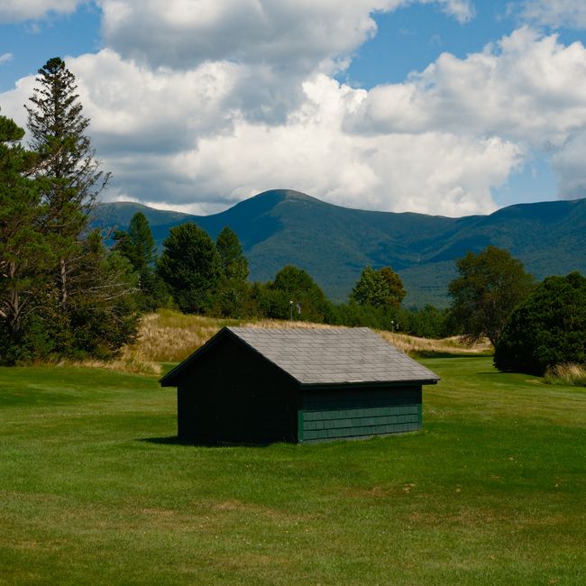 A small rustic wooden shed sits in a lush green field with a stunning mountain backdrop under a bright summer sky.