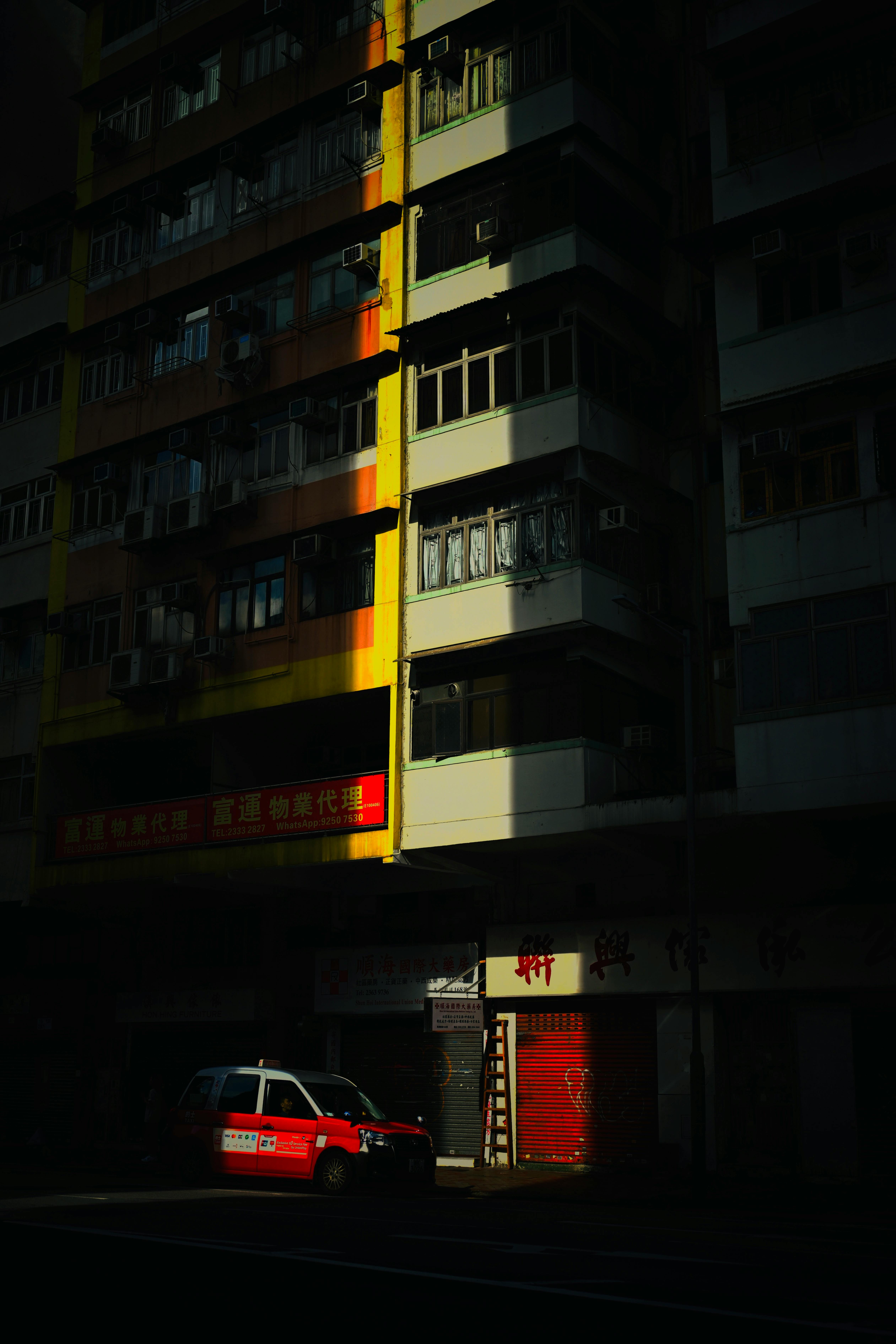 A red taxi parked in the shadow of a high-rise building during the day.