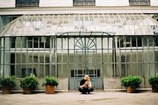 A woman sits at a modern glass atrium surrounded by potted plants.