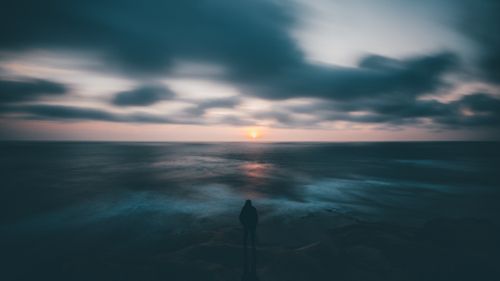 Free Silhouette of a person standing by the ocean at dusk, watching a dramatic sunset. Stock Photo
