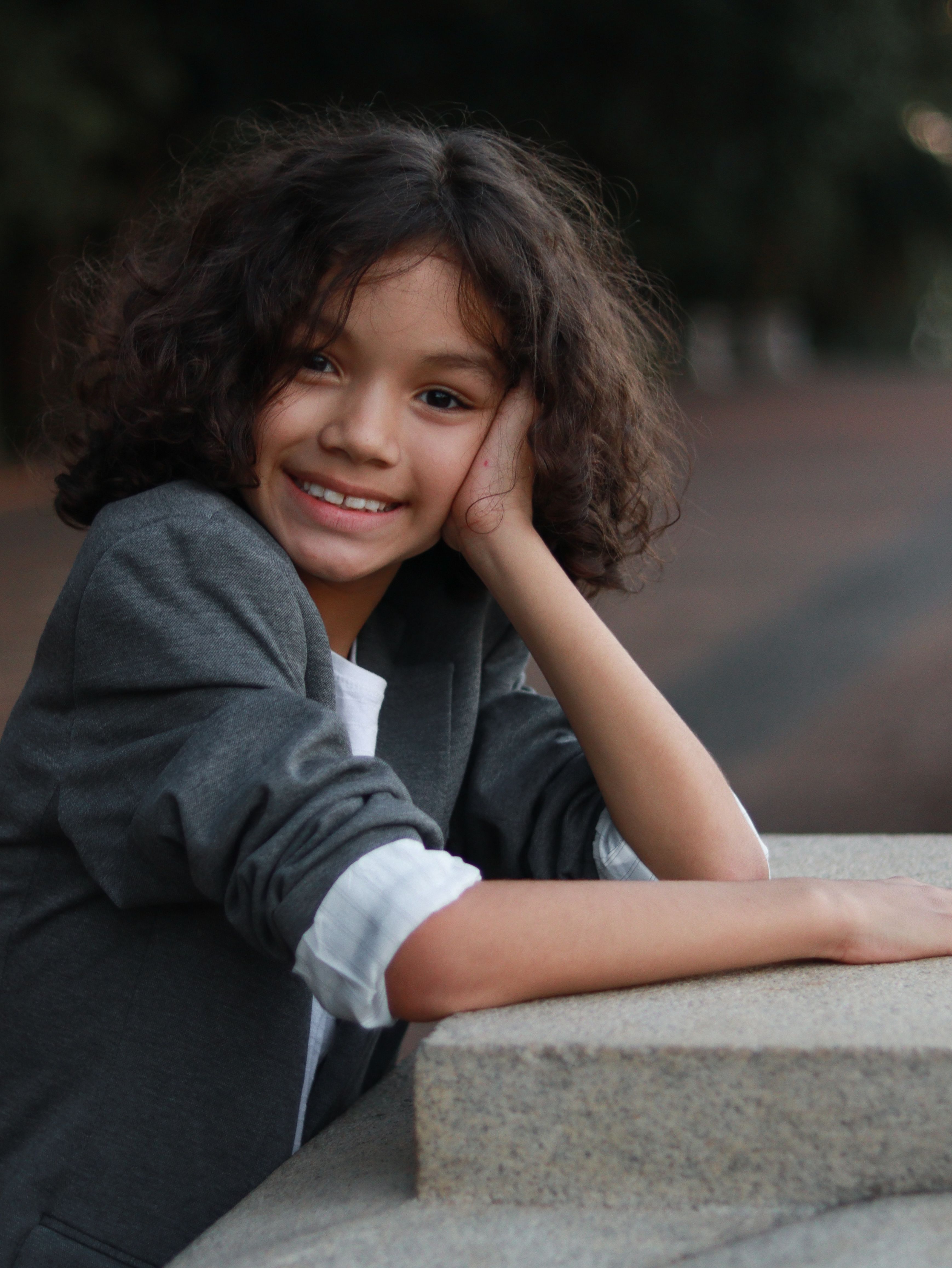A charming young girl with curly hair smiles while sitting outdoors, capturing a relaxed and joyful moment.