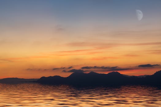 Tranquil ocean view during a vibrant sunset with distant mountains and a crescent moon.