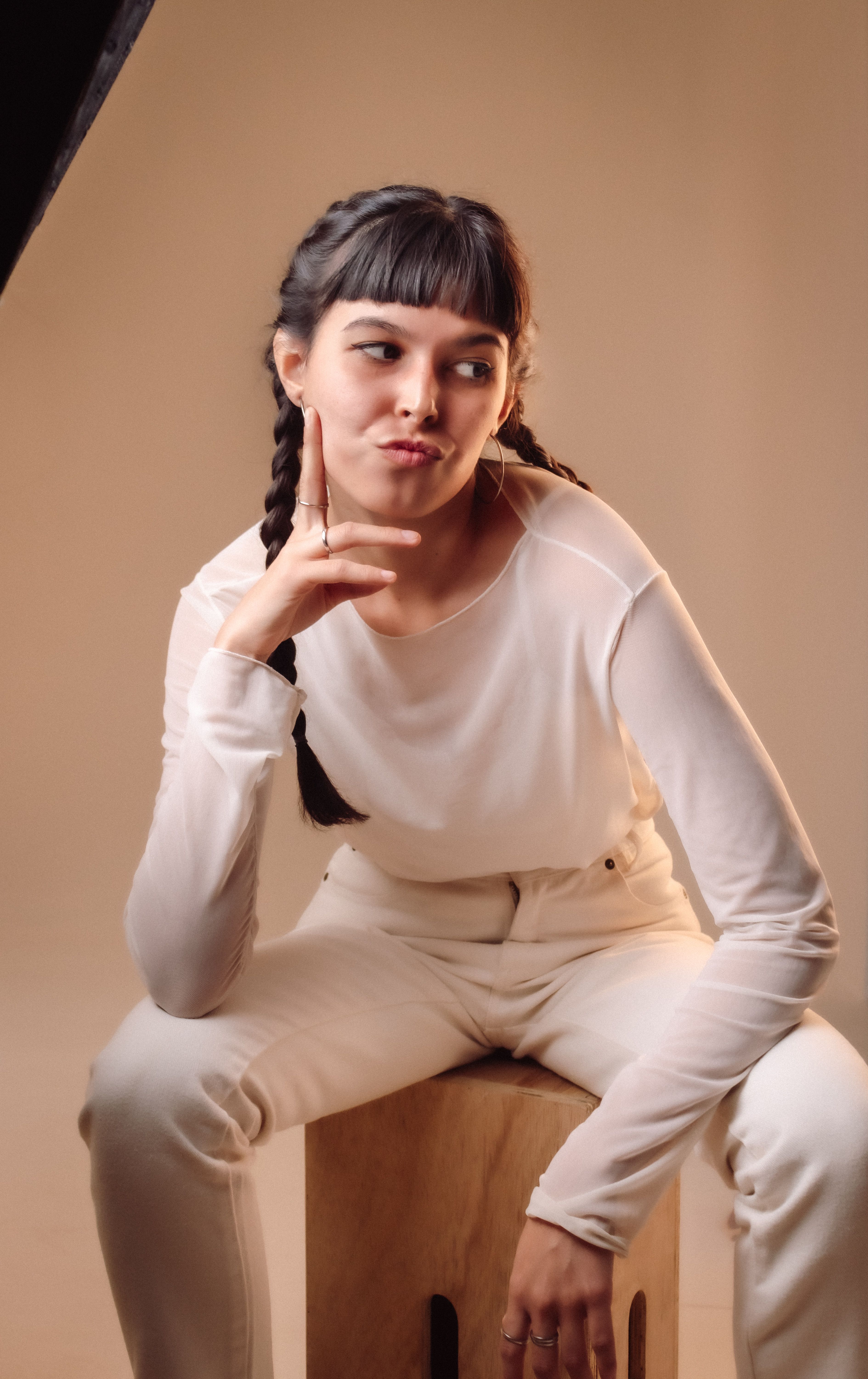A young woman with braids sitting on a stool indoors in casual white attire.