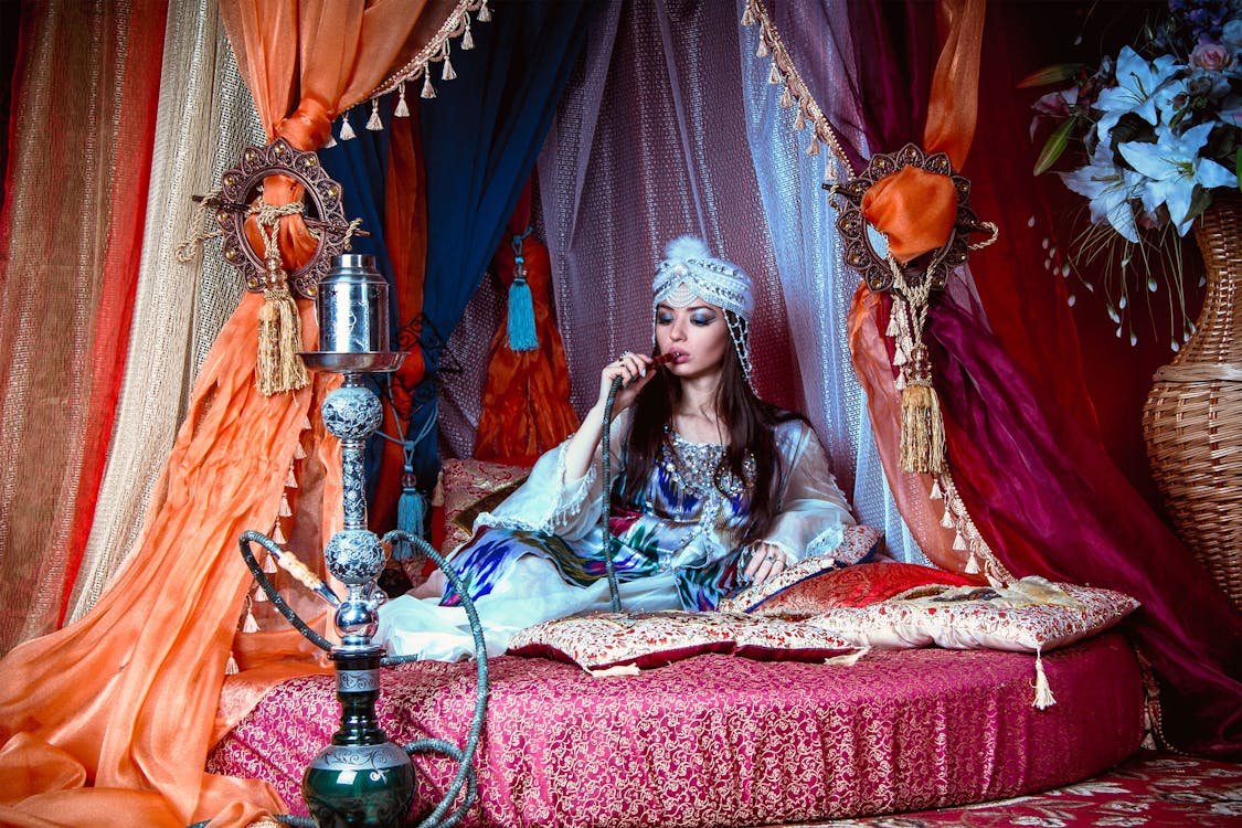 Woman Wearing White and Blue Dress Lying on Multicolored Bed and Smoking Bong