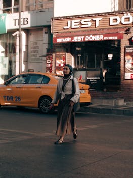 A woman walks across a city street in front of a doner shop and a yellow taxi, capturing urban life.