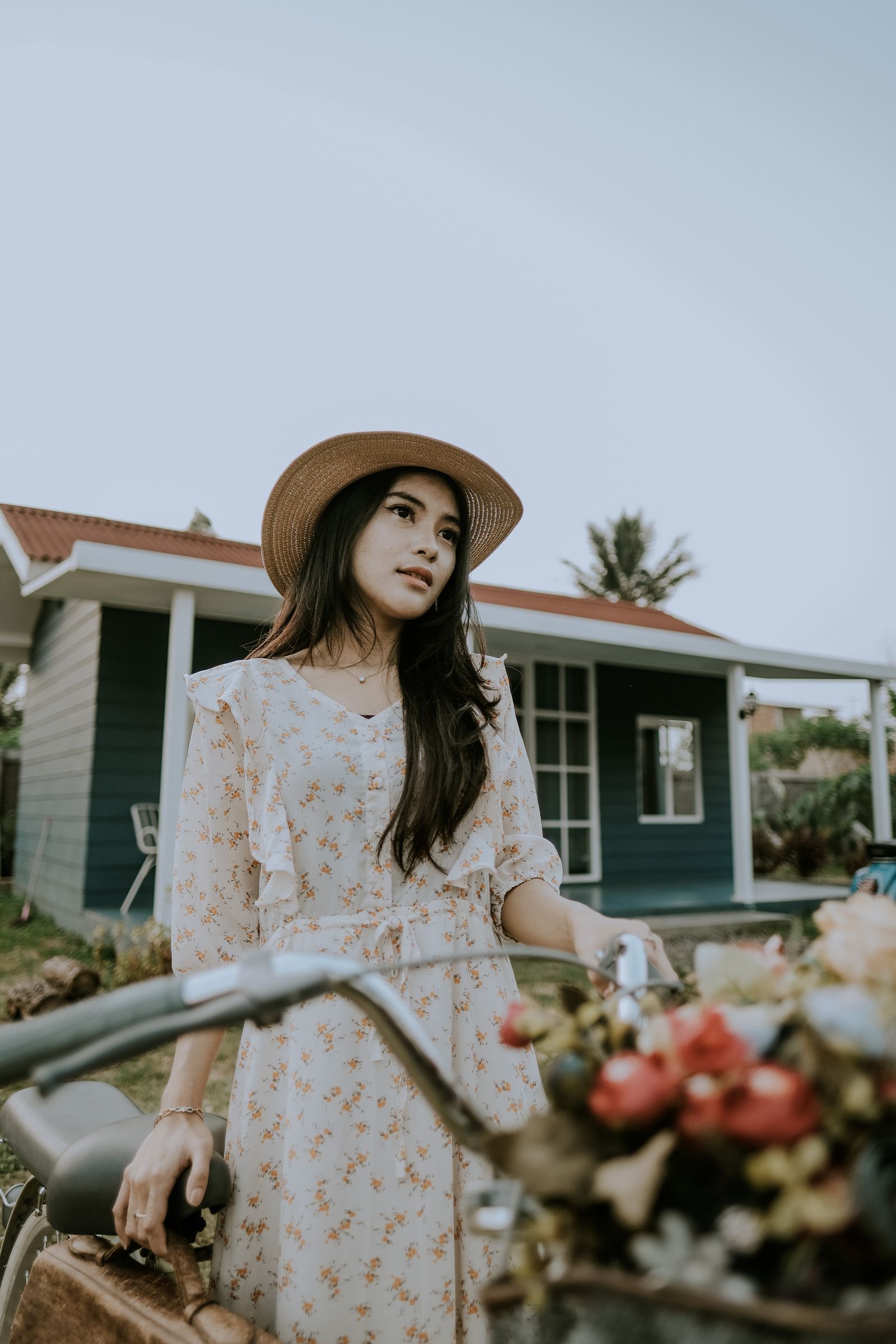 A woman in a hat and dress standing next to a bicycle