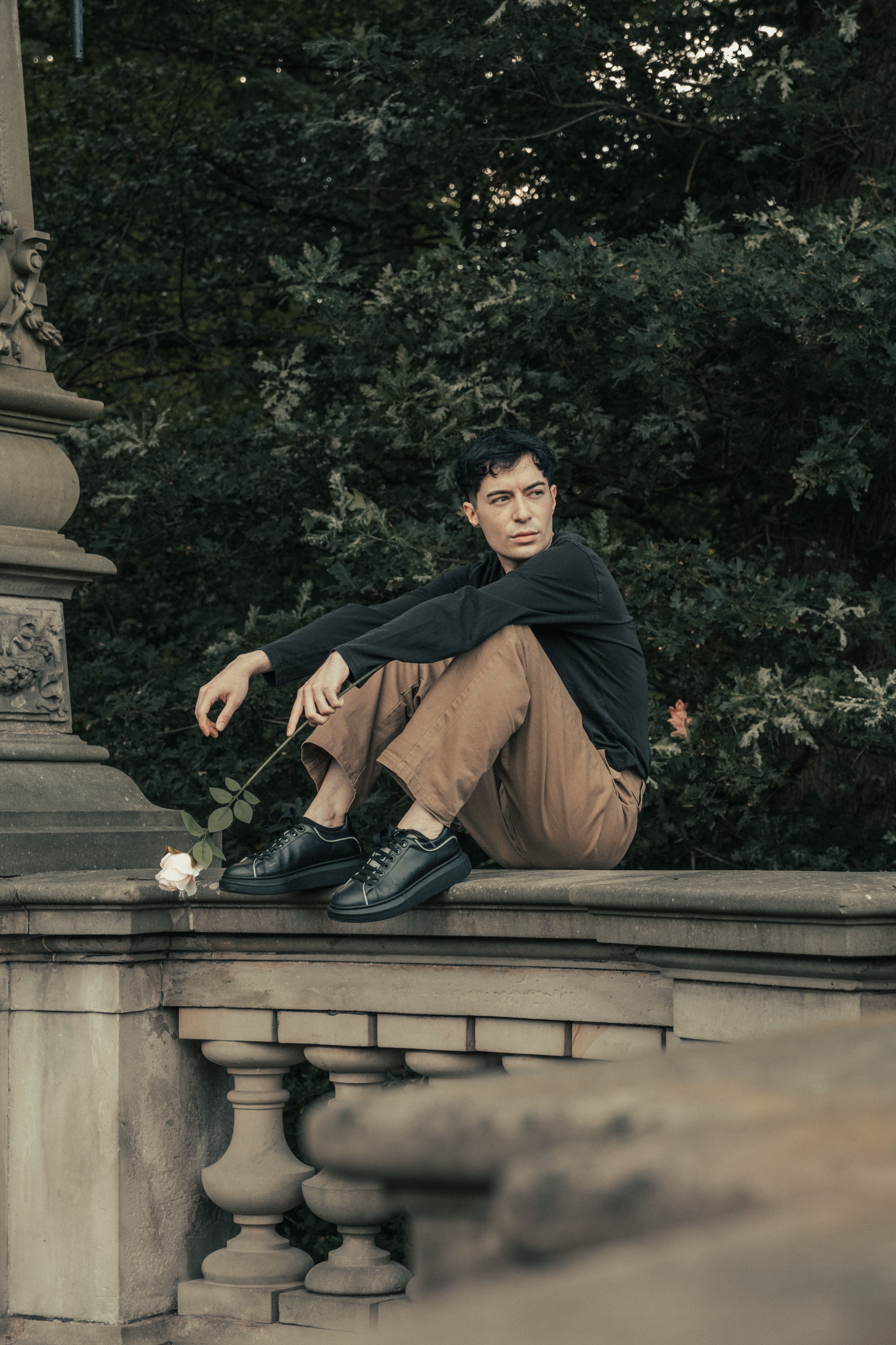 A young man sitting on a stone bench with a contemplative look, holding a rose.