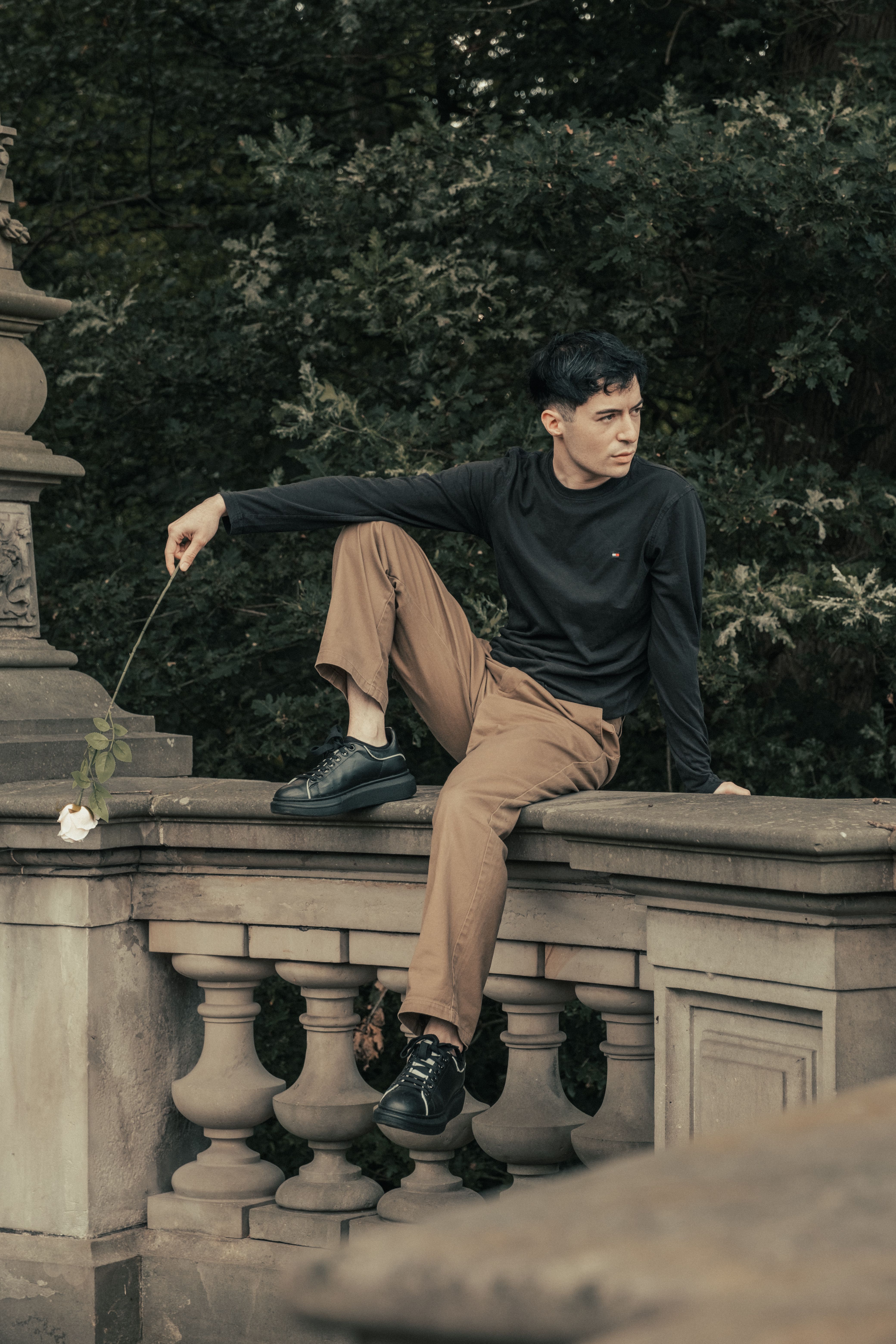 A young man sits on a stone sculpture holding a rose, outdoors in a garden setting.