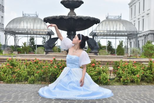 A graceful woman in a blue dress poses elegantly in an outdoor garden with fountain and classical architecture.