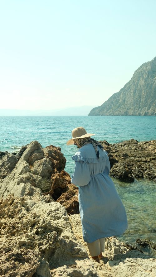 Free A woman in a summer dress and hat explores a rocky beach by the sea. Stock Photo