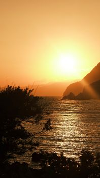 Golden sunset illuminating the sea with silhouetted cliffs and trees, creating a serene landscape.