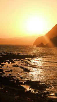 A stunning sunset over a tranquil, rocky seashore with golden hues reflecting on the water.
