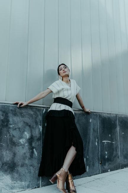 Elegant woman in contemporary attire posing against a textured wall in daylight.