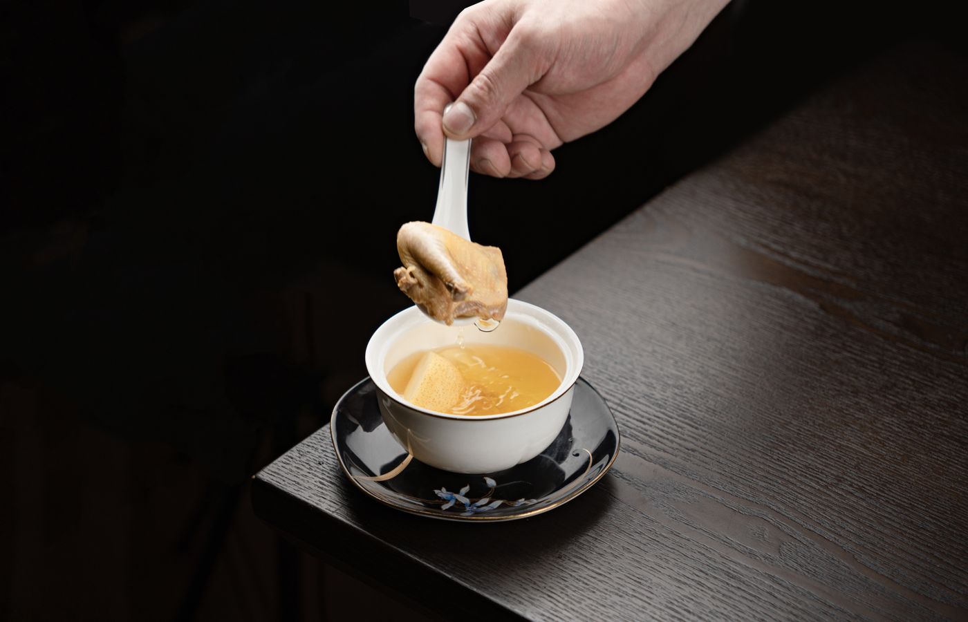 Close-up of a hand holding a spoon with chicken in a bowl of rich broth on a wooden table.