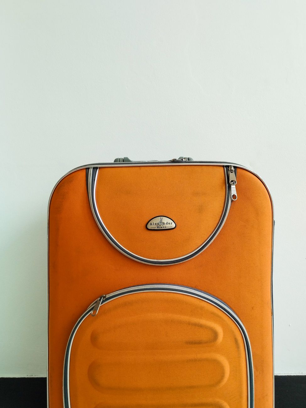 Close-up of an orange suitcase with metal accents against a plain wall.