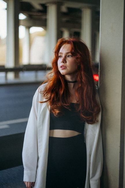 A woman with red hair and a white coat standing by a wall