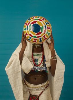 Person in traditional attire holding a vibrant, patterned hat, set against a blue backdrop.