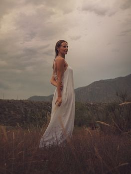 A woman in a white dress poses gracefully in the mountainous outdoors, showcasing natural beauty and elegance.