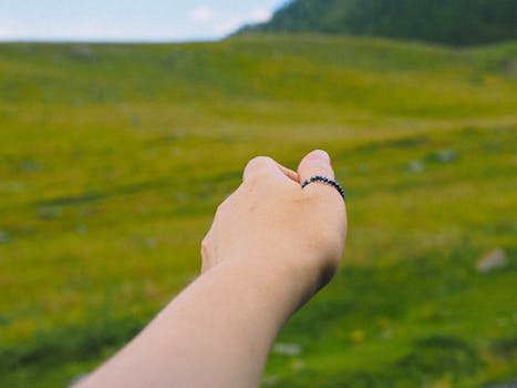 A hand with a beaded ring reaching towards a lush green landscape in summer.
