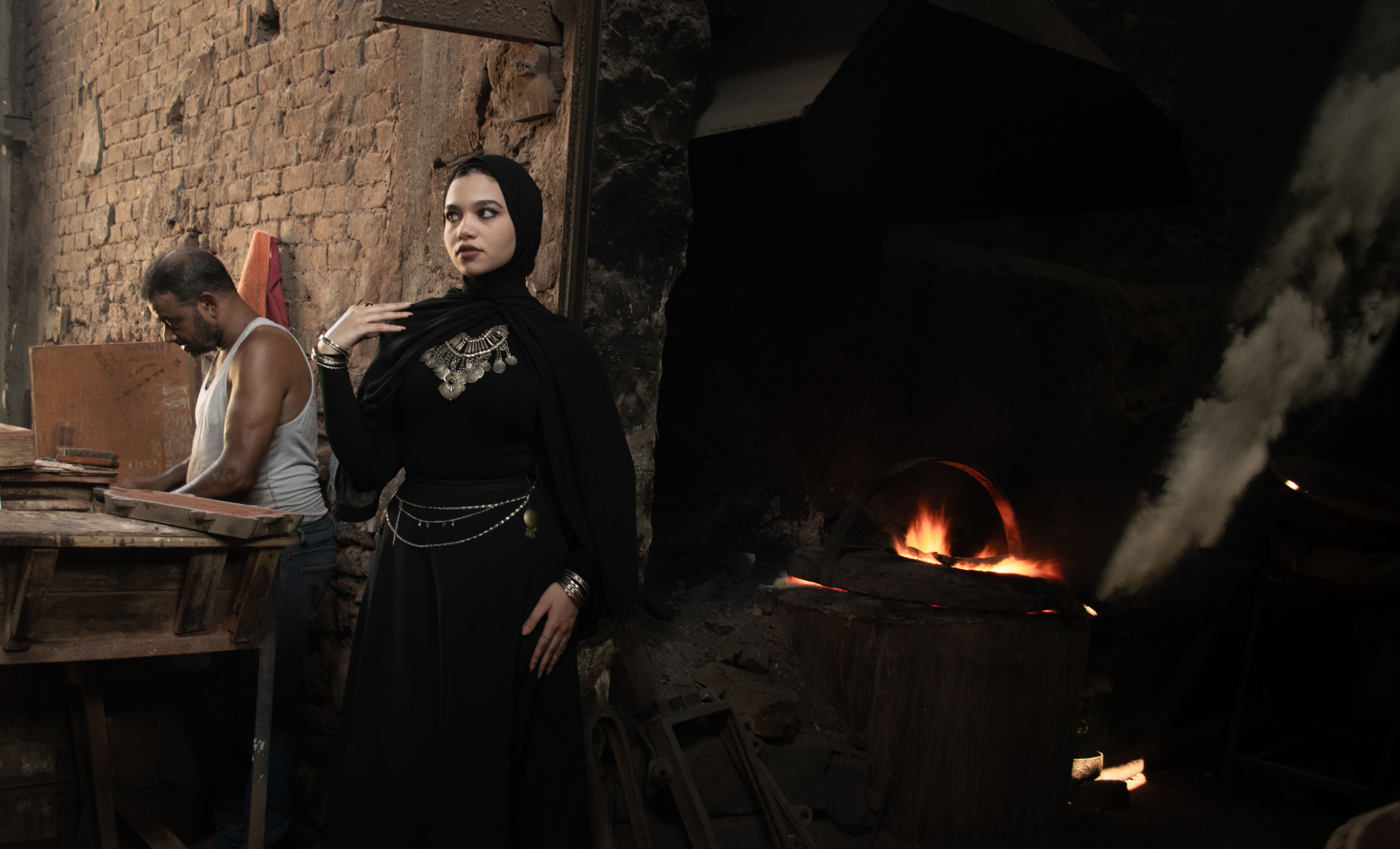 A woman in cultural attire poses in a rustic workshop with a visible flame, creating a moody ambiance.