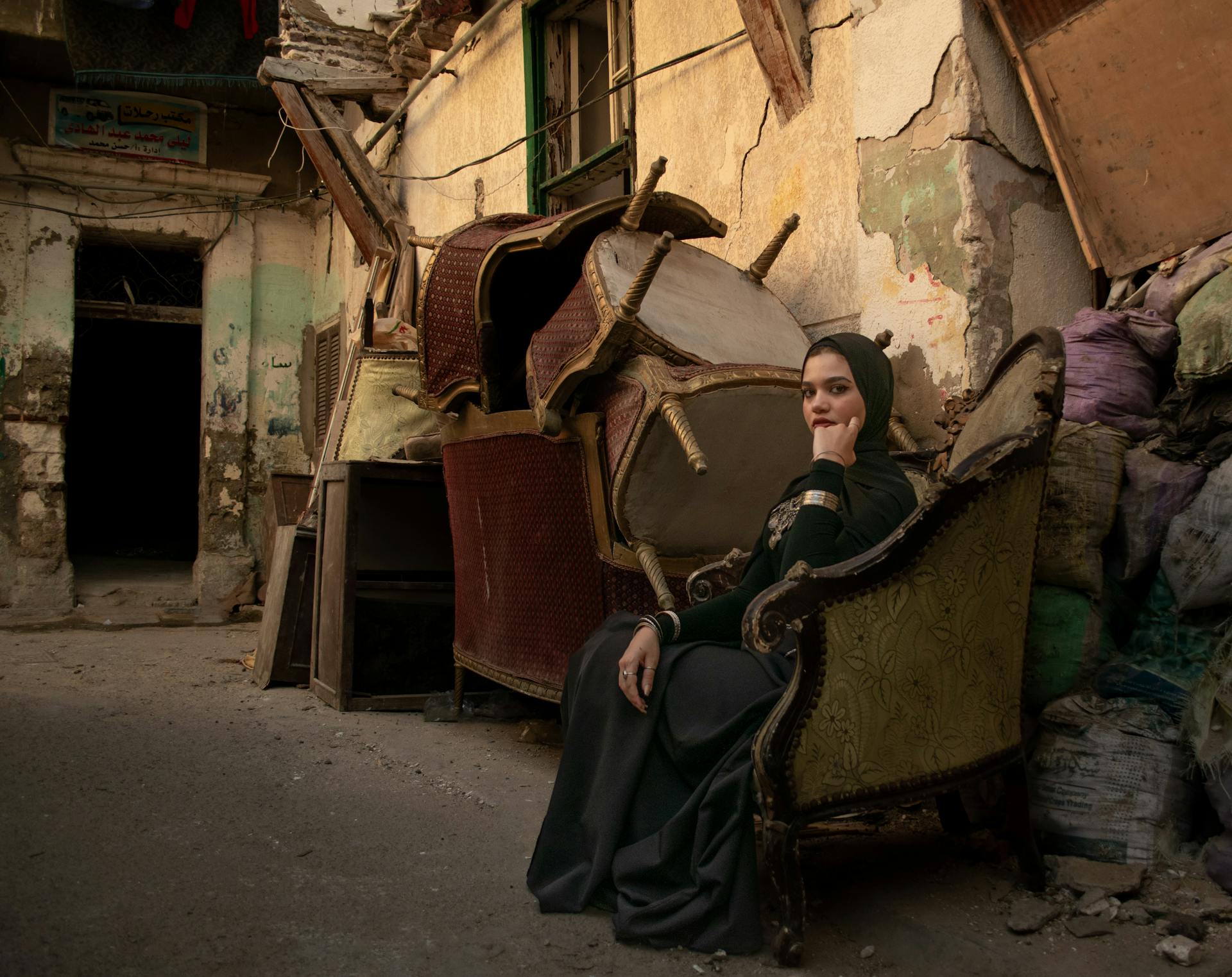 A contemplative woman in traditional attire sits amidst worn furniture and crumbling buildings.