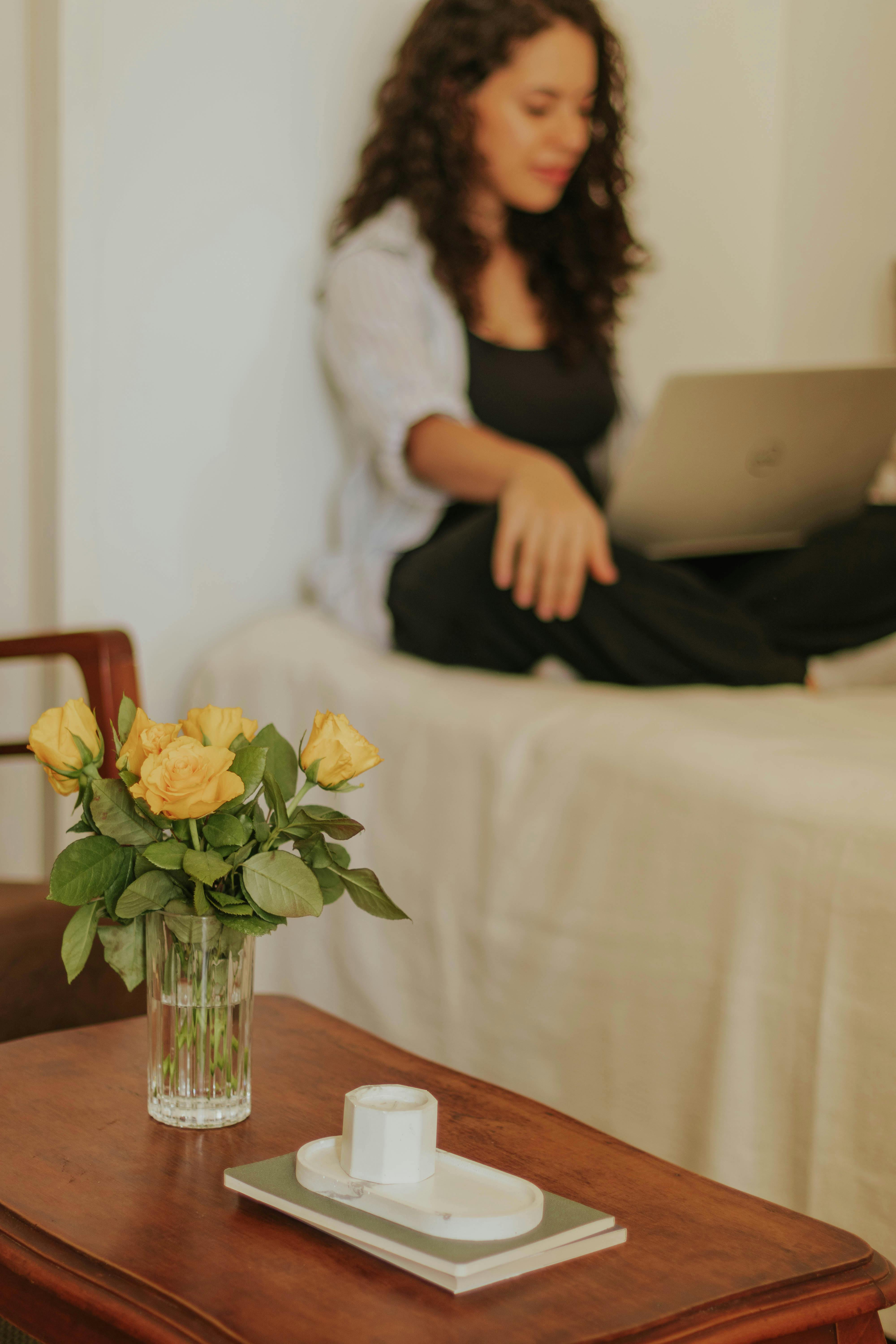 Free Young woman sitting with a laptop in a cozy room featuring flowers and a warm ambiance. Stock Photo