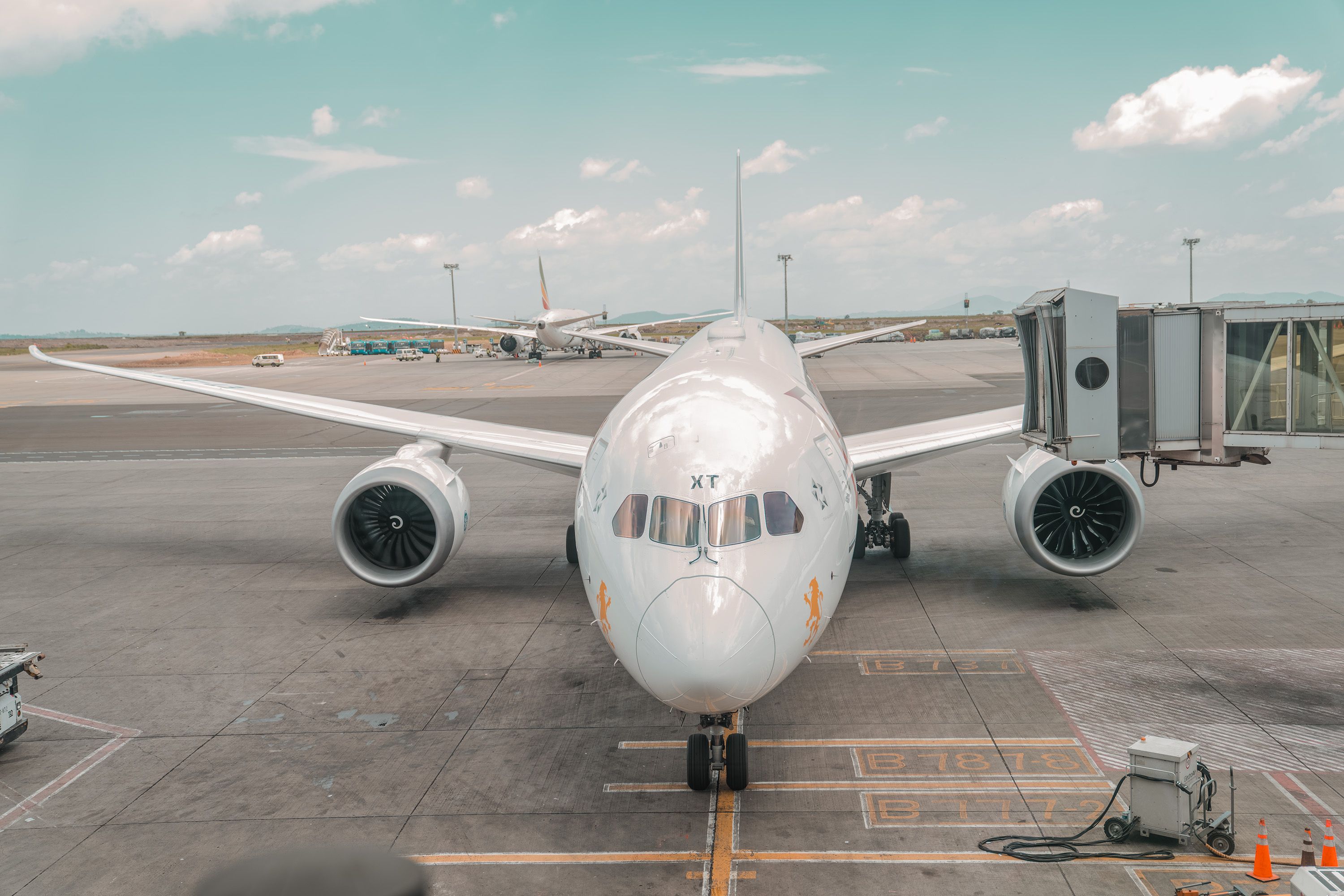 Front view of a passenger aircraft parked at an airport terminal, with a jet bridge, ground equipment and another plane behind it
