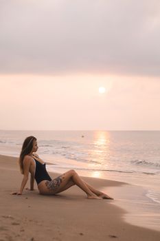 A woman in a swimsuit sits by the ocean at sunset, embracing tranquility.