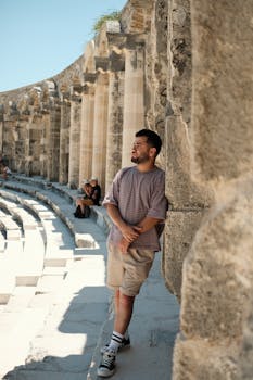 A young man leans against stone columns in an ancient amphitheater under a sunny sky.
