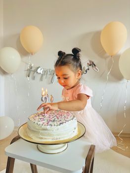 Adorable young girl in a pink dress celebrating birthday with cake and balloons indoors.