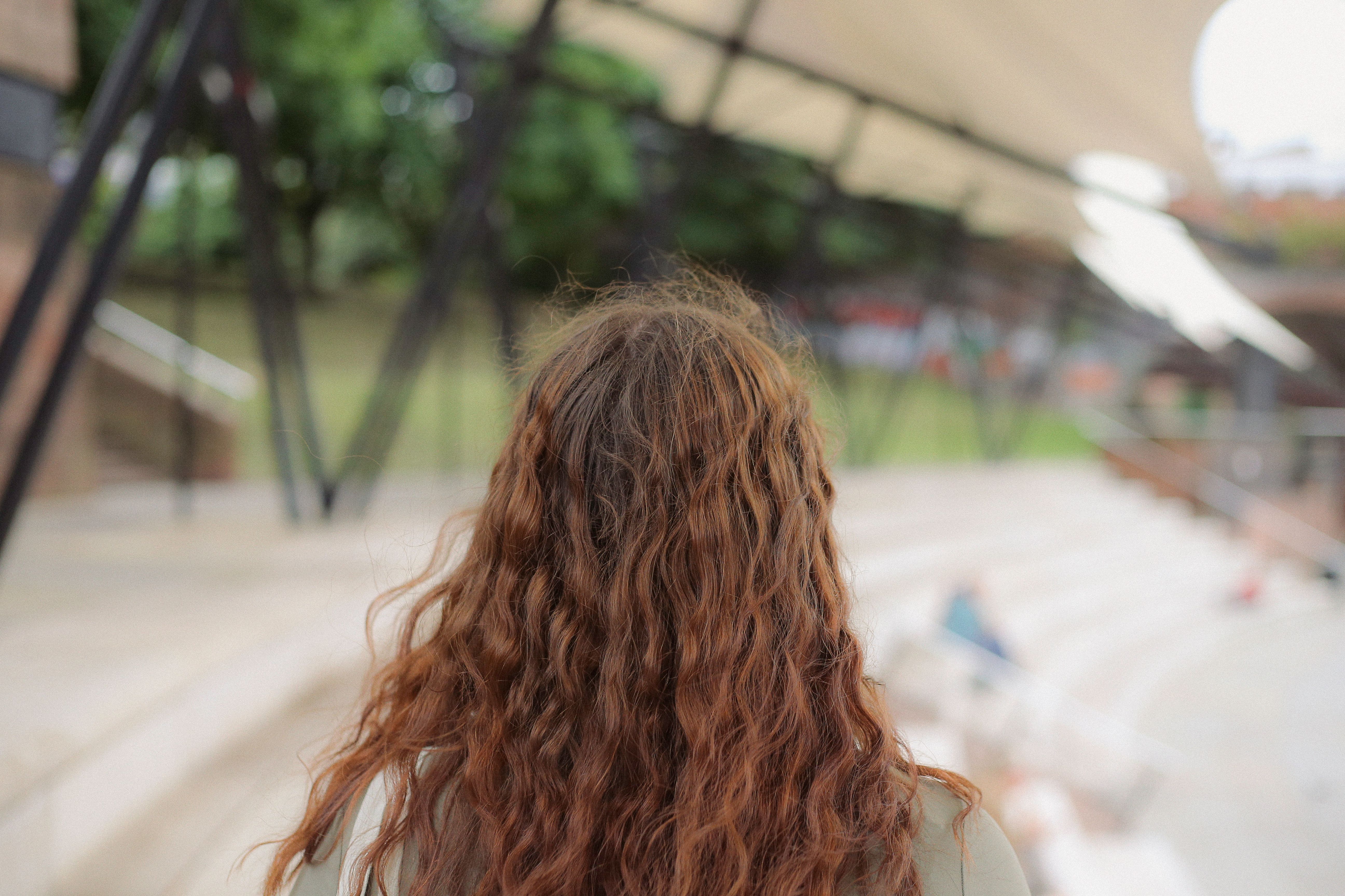 A woman with curly hair stands in an outdoor urban area, surrounded by greenery.
