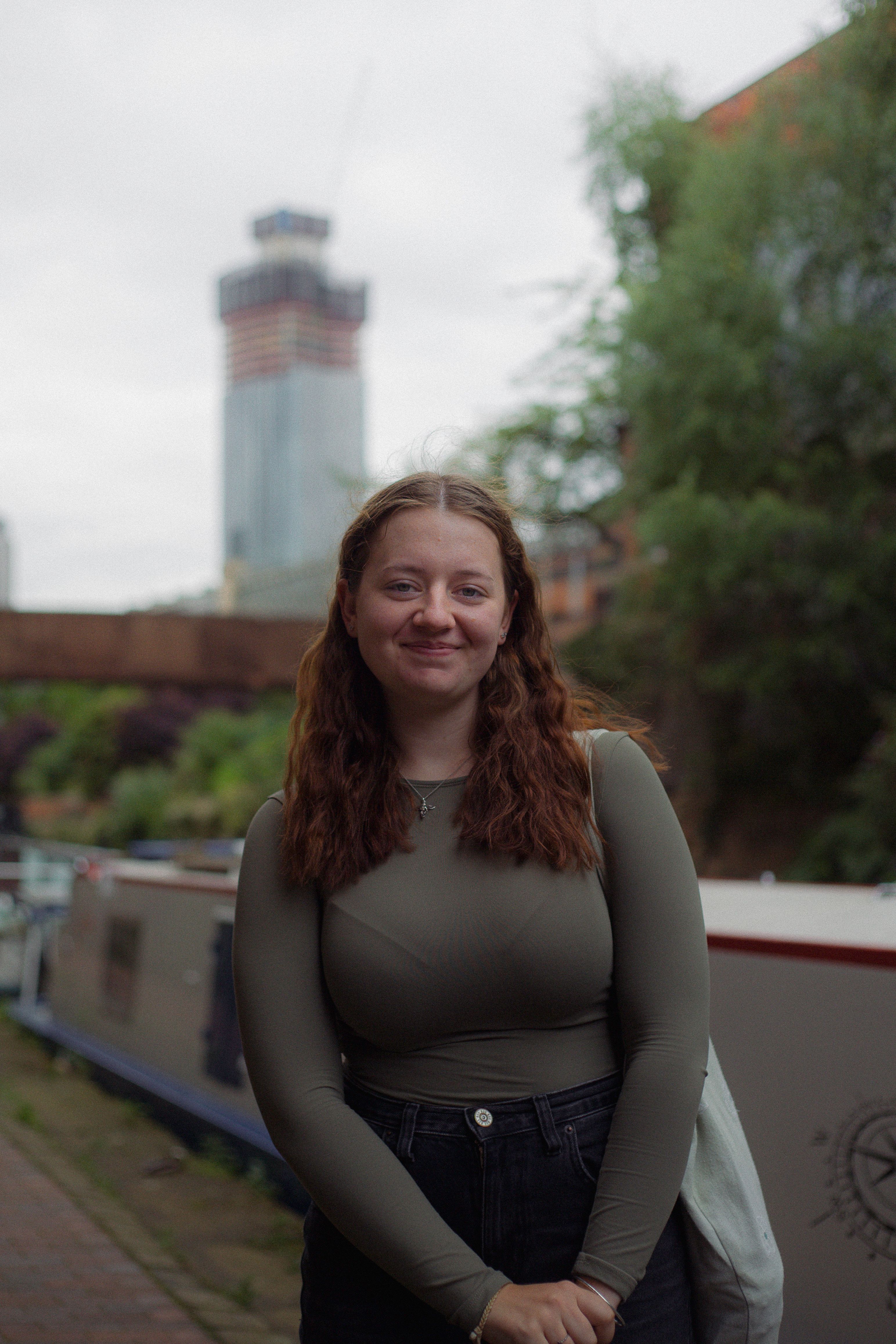 A young woman stands smiling by a canal in an urban setting on a cloudy day.
