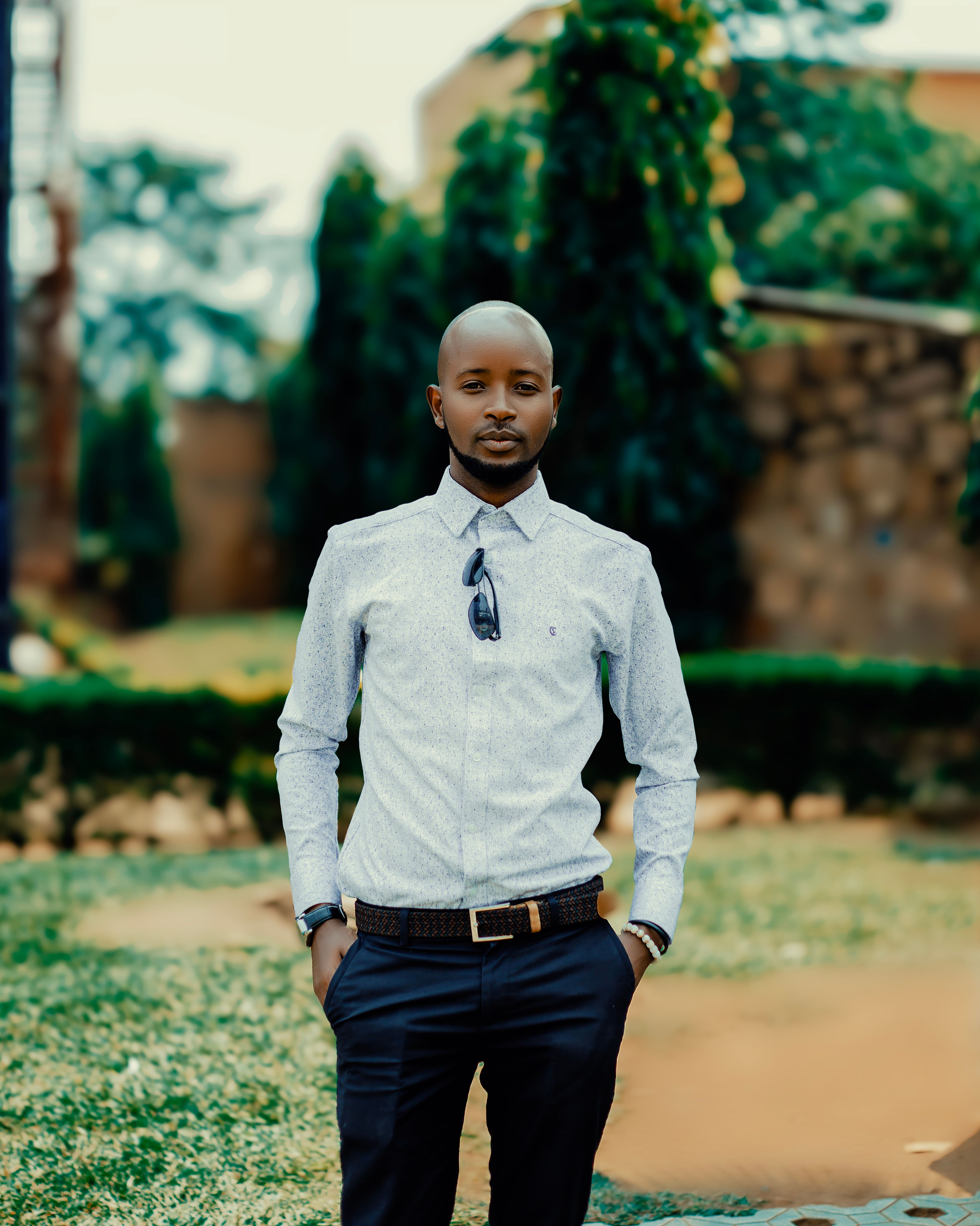 A fashionable man stands confidently outdoors with greenery in the background in Bujumbura.