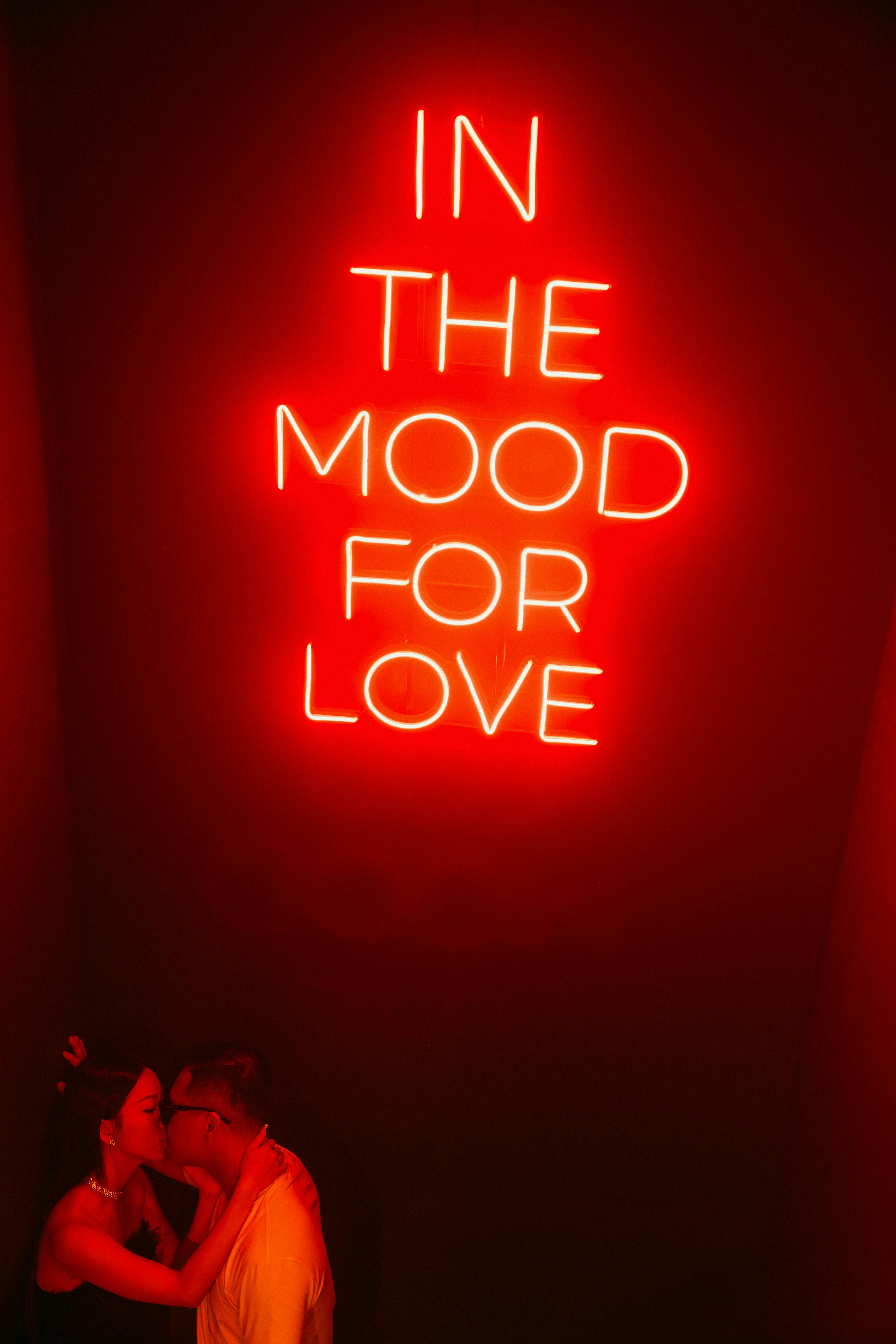 Couple embracing under a bright romantic neon sign reading 'IN THE MOOD FOR LOVE'.