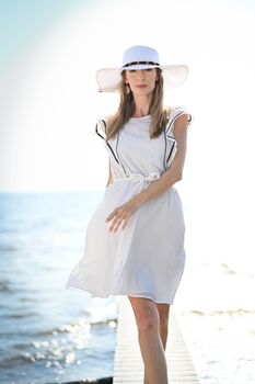 Woman in white dress and hat strolls on a sunny beach pier, exuding elegance and summer vibes.