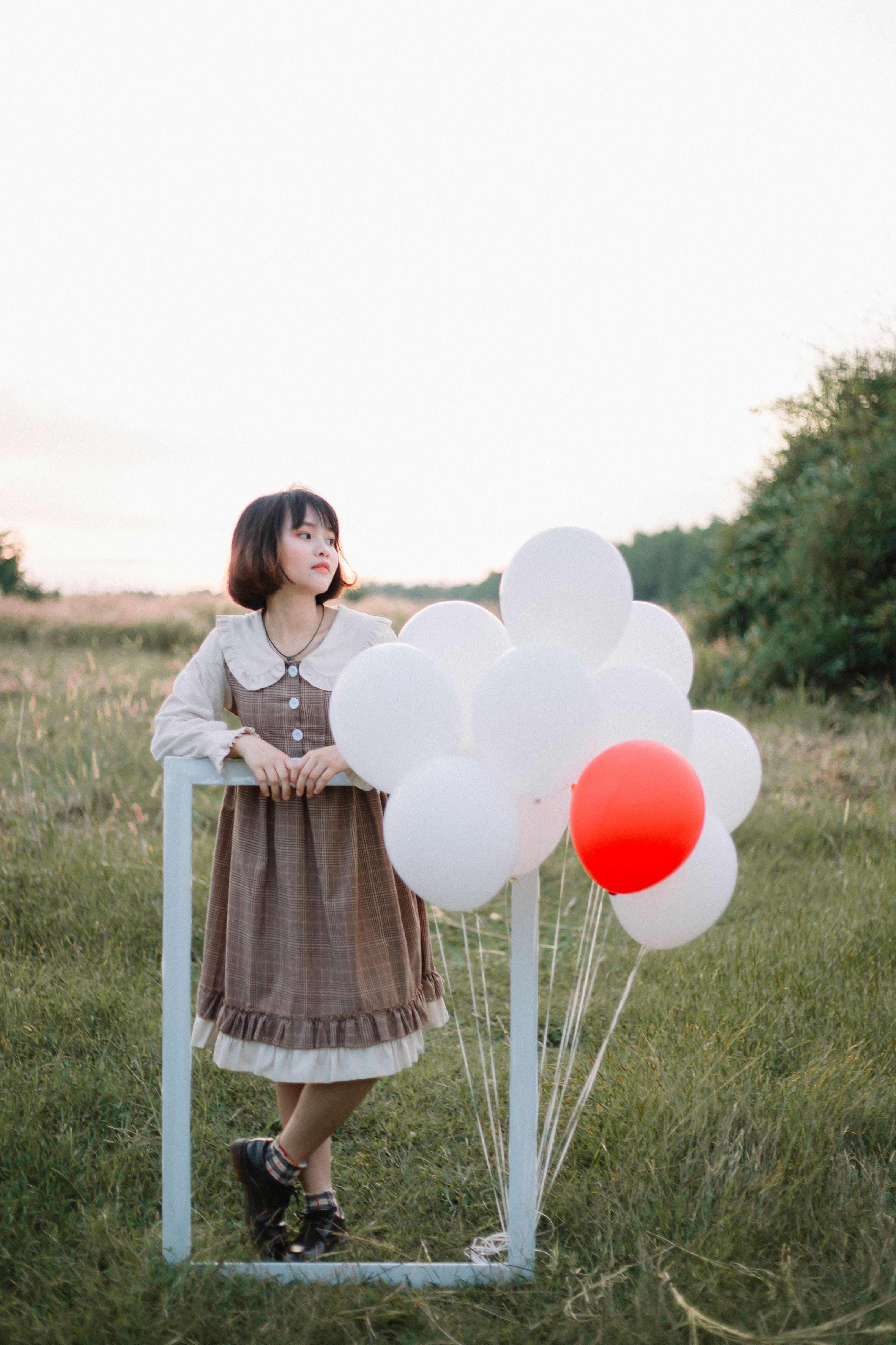 A fashionable woman in a rustic dress stands with a balloon bouquet in a serene grassy field.