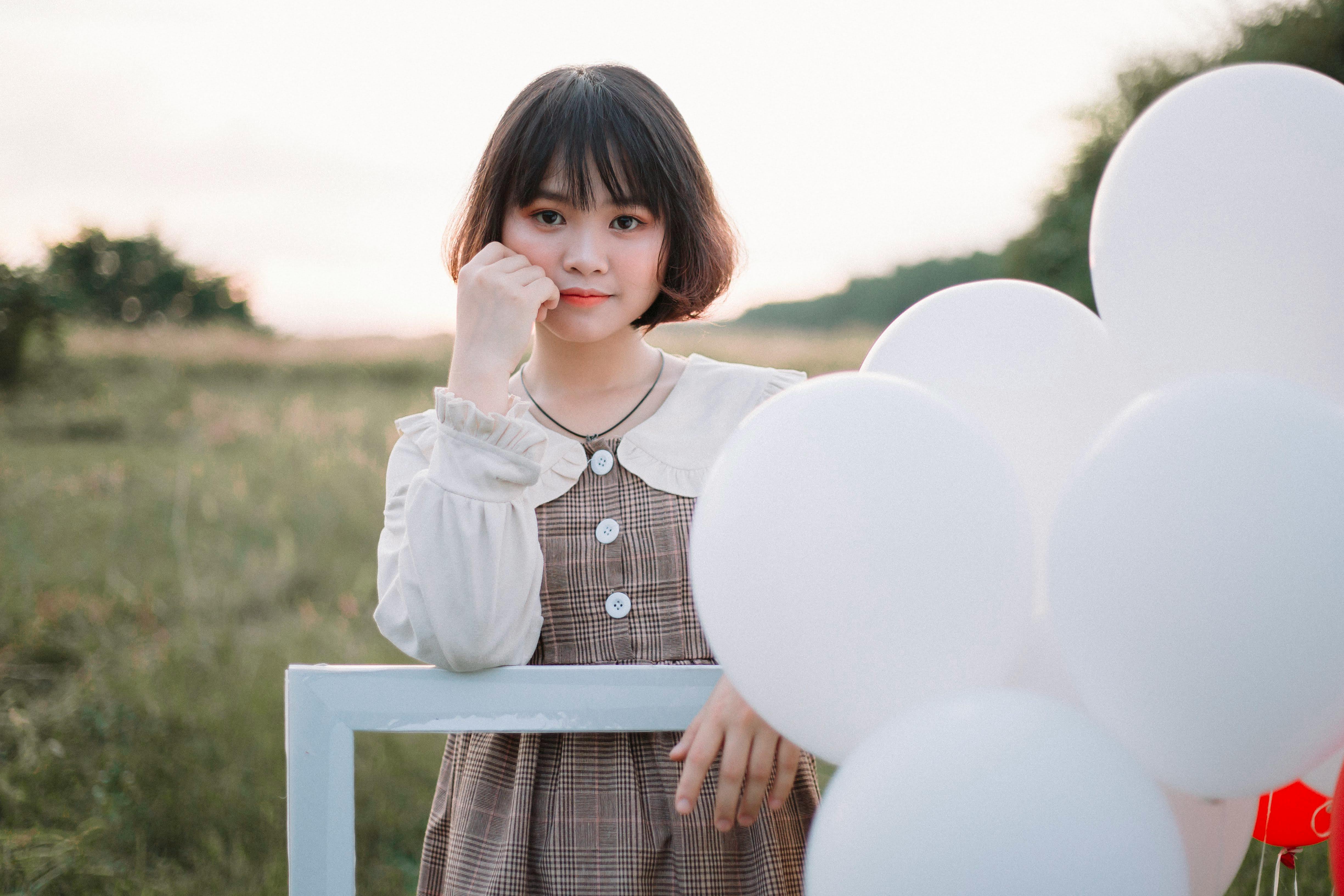 A young woman in vintage attire stands with balloons in a scenic field, evoking a whimsical vintage charm.