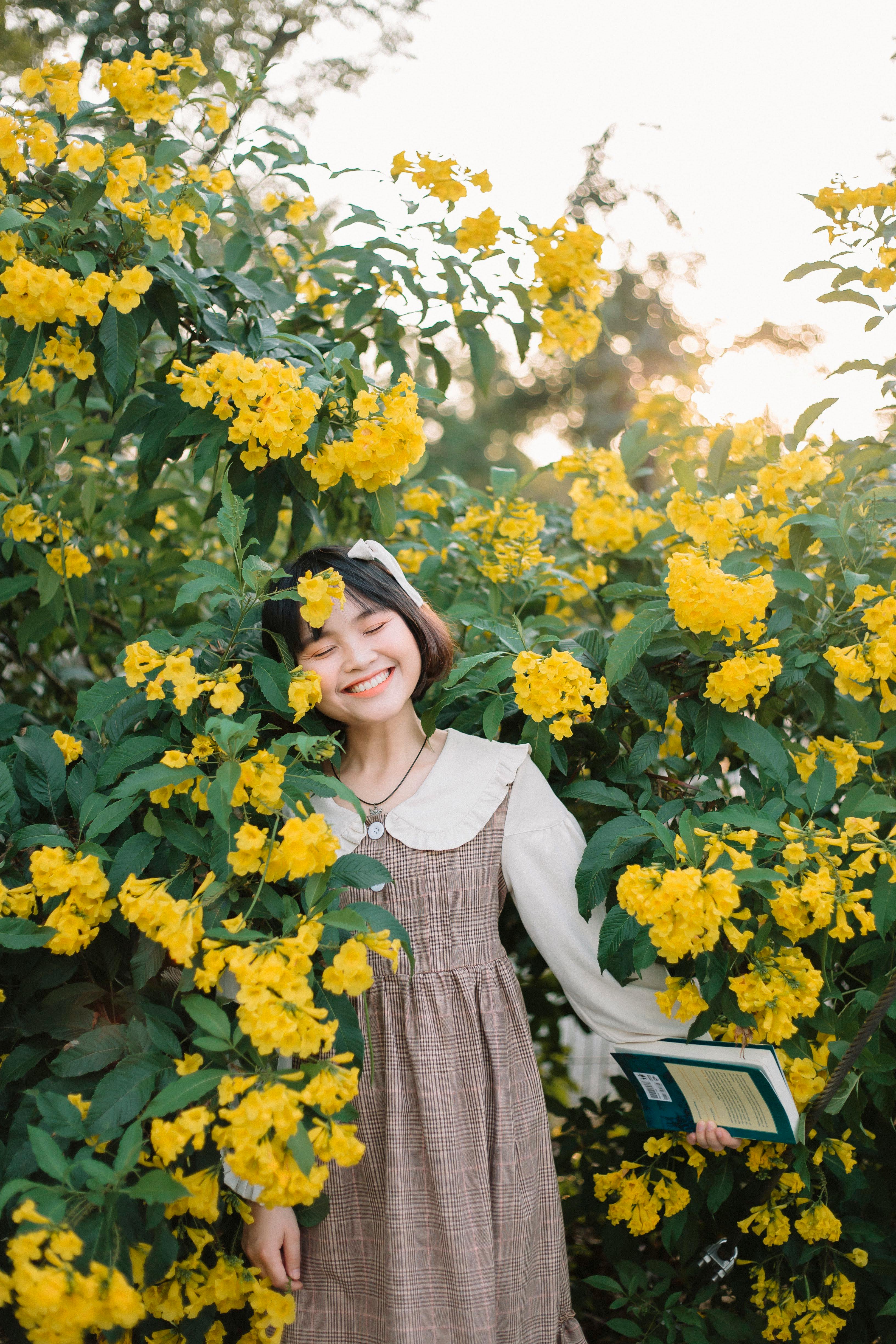 Smiling woman in a floral dress stands among vibrant yellow flowers, eyes closed in bliss.
