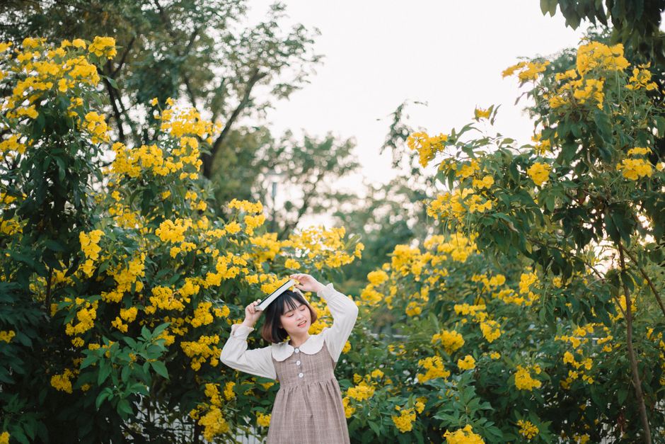 A young woman stands amid vibrant yellow flowers holding a book above her head, evoking a playful moment outdoors.