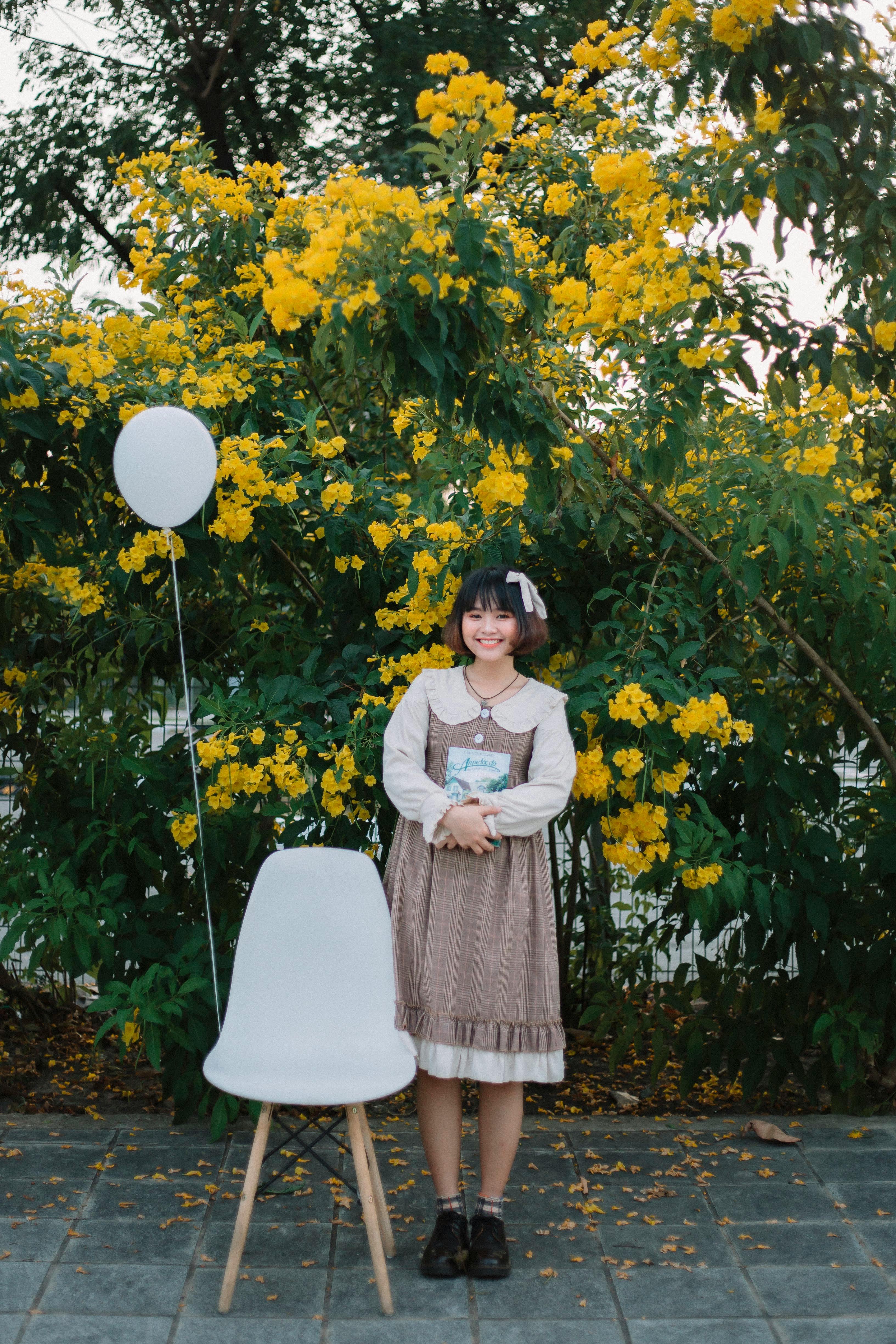 Asian woman in vintage dress smiling with a book beside yellow flowers and a balloon.