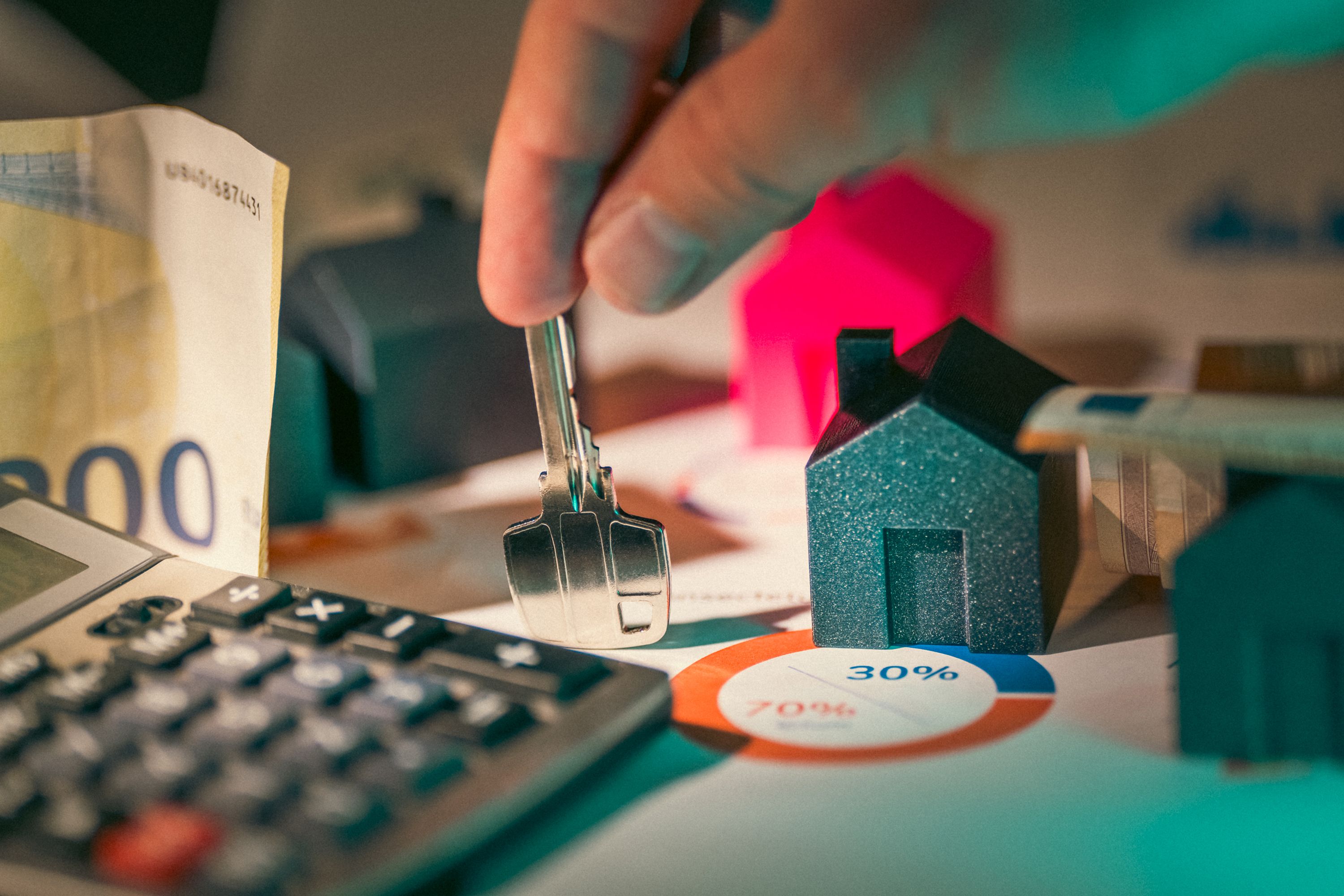 Alt Text: Close-up of a person's hand holding a silver house key over a desk with small model homes, banknotes, a calculator, and printed financial pie charts.