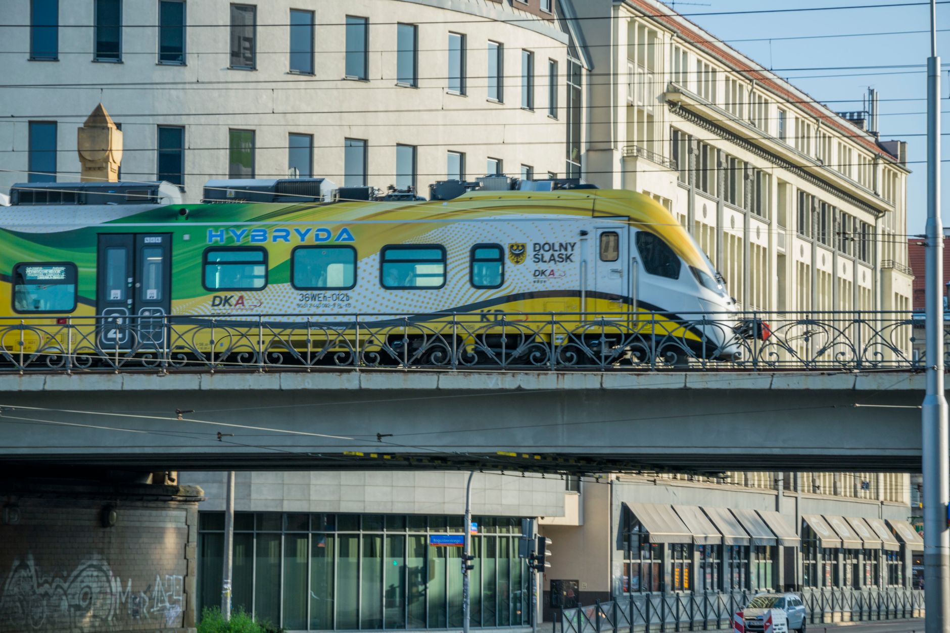 A hybrid train travels on an elevated railway in Wrocław, Poland, amidst urban architecture.