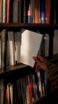 A person pulls out a blank white book from a shelf filled with various colored books.