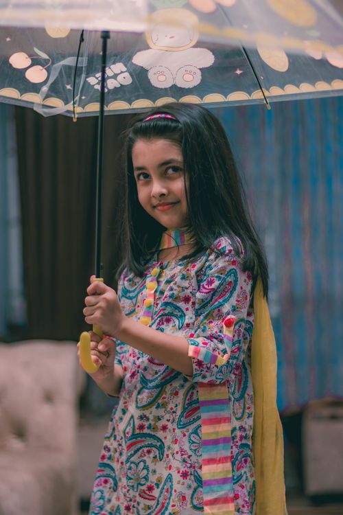 Free Charming young girl with a colorful outfit smiling and holding a whimsical umbrella indoors. Stock Photo