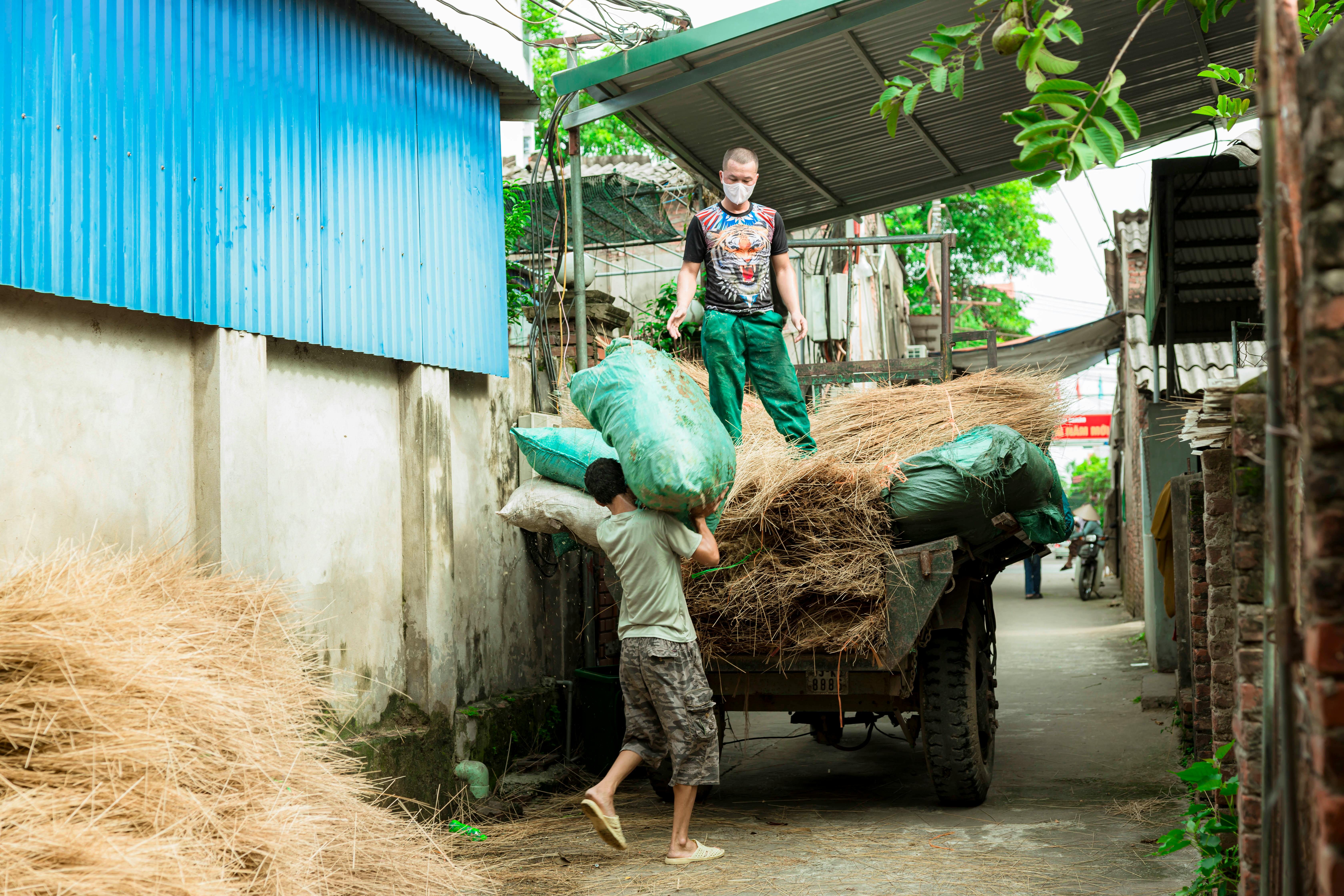 Two men load straw bundles onto a cart in a rural alley, working hard on a bright day.