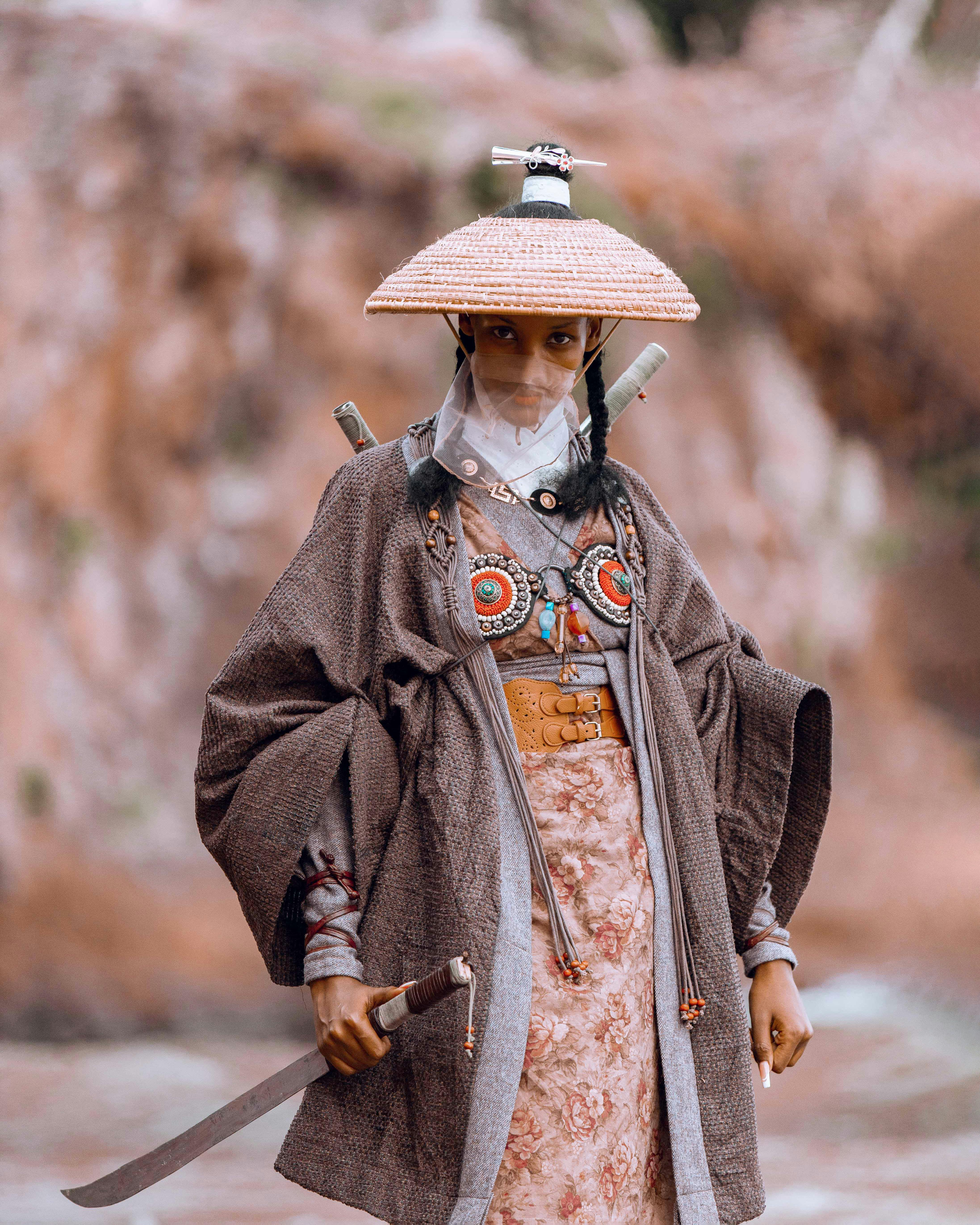 A traditionally dressed warrior stands confidently outdoors, showcasing intricate attire and a sword.