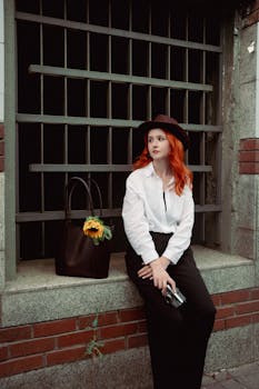Fashionable redheaded woman sits outdoors with a camera and bag, exuding vintage style.