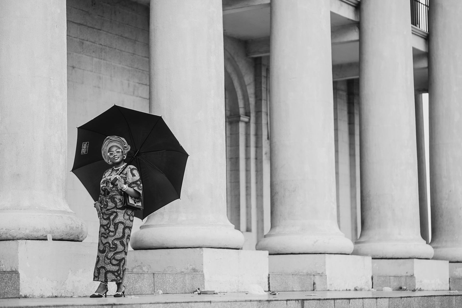 An African woman in traditional attire poses with an umbrella beside classic architectural columns in Ibadan.